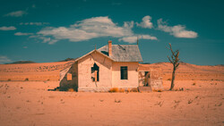 Abandoned house with peeling pink walls and dark windows stands alone in vast, arid desert landscape.