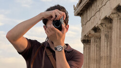 Photographer holding a rangefinder camera to his eye while standing in front of classical stone columns.