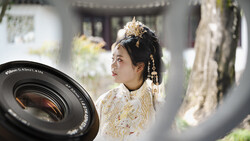 Woman in ornate white and gold traditional dress with elaborate gold headdress, photographed through a camera lens in soft natural light.