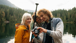 Three people holding a paddle while standing in a boat on a calm alpine lake surrounded by forested mountains
