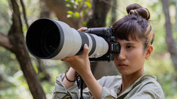 Young woman holding a telephoto lens to her eye in a wooded outdoor setting