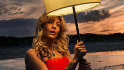 Woman in red strapless dress holding illuminated lamp shade against dramatic sunset over water