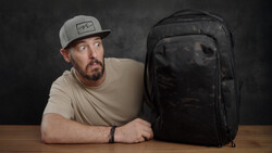 Man in tan cap and beige shirt posing with black backpack at wooden table