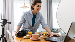 Female content creator at wooden desk with professional DSLR camera, ring light, and laptop during food photography setup