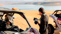 Photographer with telephoto lens shooting beside off-road vehicles overlooking desert reservoir landscape