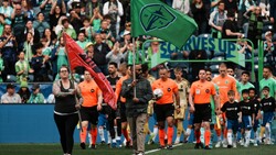 Soccer team in orange jerseys walks onto field during pre-game ceremony with flag bearer and spectators in background