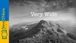 Black and white mountain landscape with a prominent peaked ridge and distant peaks under dramatic cloudy sky.