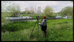 Photographer operating a surveying theodolite on tripod in green field with industrial cooling towers in background