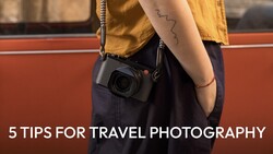 Photographer wearing yellow shirt and dark pants holding compact camera with chain strap against rust-colored wall