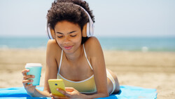 Woman wearing headphones lying on beach towel holding smartphone and beverage