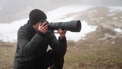 Photographer in black winter gear crouching while composing shot with telephoto lens in snowy mountain terrain