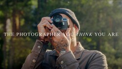 Photographer with tattooed arm holding vintage film camera to face, wearing beanie and jacket in forested setting