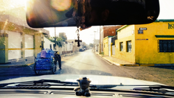 Dashboard view through a vehicle windshield looking down a colorful street with blue cargo tricycle and yellow buildings