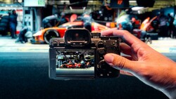 Hand holding mirrorless camera displaying racing pit crew on LCD screen with blurred race car in background