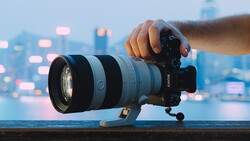 Hand holding Sony mirrorless camera with telephoto zoom lens mounted on tripod head, city skyline with bokeh lights in background