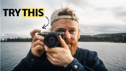 Photographer holding rangefinder camera toward viewer with lake and forested mountains in background