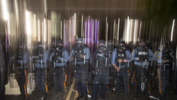 Row of riot police in tactical gear holding shields and batons, illuminated by vertical neon light tubes