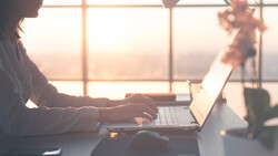 Woman working on laptop at desk by sunlit window during golden hour