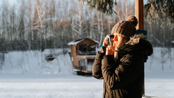 Person in winter coat and knit hat holding warm beverage outdoors in snowy landscape