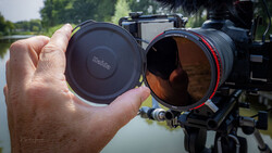 Photographer holding lens cap andND filter in front of mounted cinema camera overlooking water