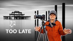 Photographer with camera on tripod wearing orange jacket, with pier structure and text overlay reading "TOO LATE"