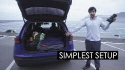 Filmmaker holding camera beside open car trunk packed with gear overlooking coastal highway