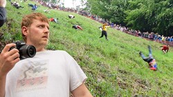 Photographer with camera documenting competitors rolling down grass-covered hill during outdoor event