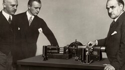 Three men in business suits examine a large industrial machine on a table