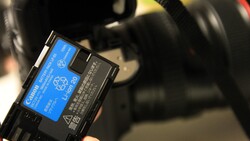 Hand holding a blue Canon LP-E6 lithium-ion battery pack in front of a blurred DSLR camera and lens.
