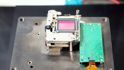 Disassembled camera internals on a metal plate: a metal-framed module with a pink-tinted sensor beside a green circuit board