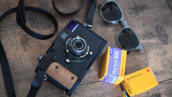 Vintage rangefinder camera with leather strap, sunglasses, and film boxes on wooden surface