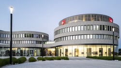 Curved modern building at dusk with red Leica logo signs on its upper level and an illuminated glass ground floor.