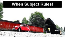 Red vintage Citroën 2CV parked against red corrugated metal wall with large dark sculptural figure looming above