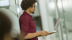 Man in maroon polo shirt holding open laptop, standing in modern office corridor with glass partitions
