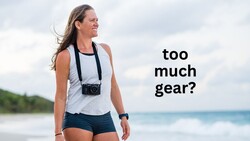 Female photographer in casual beachwear holding a camera on a strap, looking toward the ocean with text asking 'too much gear?'