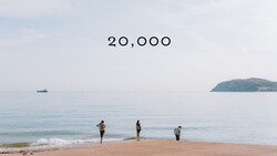 Four figures standing in shallow ocean water on a sandy beach with calm sea and distant island