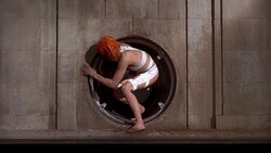 Person with orange hair curled in fetal position inside circular opening in wooden wall