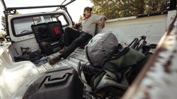 Man sitting in open truck bed with equipment and gear visible during daytime shoot