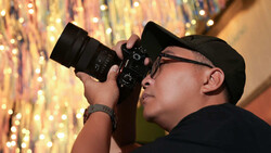 Photographer in black cap and glasses holding DSLR camera, shooting toward warm-toned bokeh backdrop