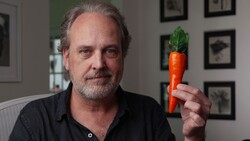 Man holding a fresh carrot with green leafy top in an indoor studio setting