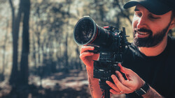 Bearded cinematographer in cap holding cinema camera with telephoto lens in forest
