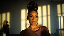 Woman with hair in a high bun and sculptural wire hoop earrings, lit by warm sunlight streaming through windows onto a wall behind her.