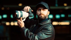Bearded photographer holding a telephoto lens and mirrorless camera against blurred stadium lights