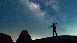 Silhouette of a person carrying a tripod over one shoulder, standing on a rock ridge beneath a bright Milky Way sky.