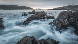 Long exposure of silky surf flowing around dark, jagged rocks on a coastline under a moody overcast sky