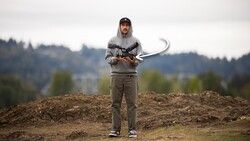 Man in a gray hoodie and cap holding a video camera rig with a top-mounted shotgun microphone on a dirt mound outdoors