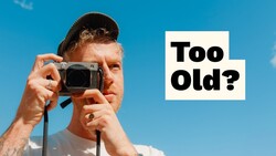 Man holding compact digital camera to his face against blue sky with "Too Old?" text overlay