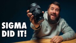 Bearded man holding a Sigma camera lens toward the camera against a dark blue background