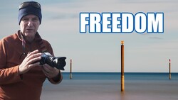Photographer holding a camera on a beach with wooden pilings and calm water, with text overlay reading 'FREEDOM'