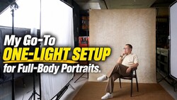 Man seated on a wooden chair in a studio, posed against a textured beige backdrop with a softbox on a stand at left.